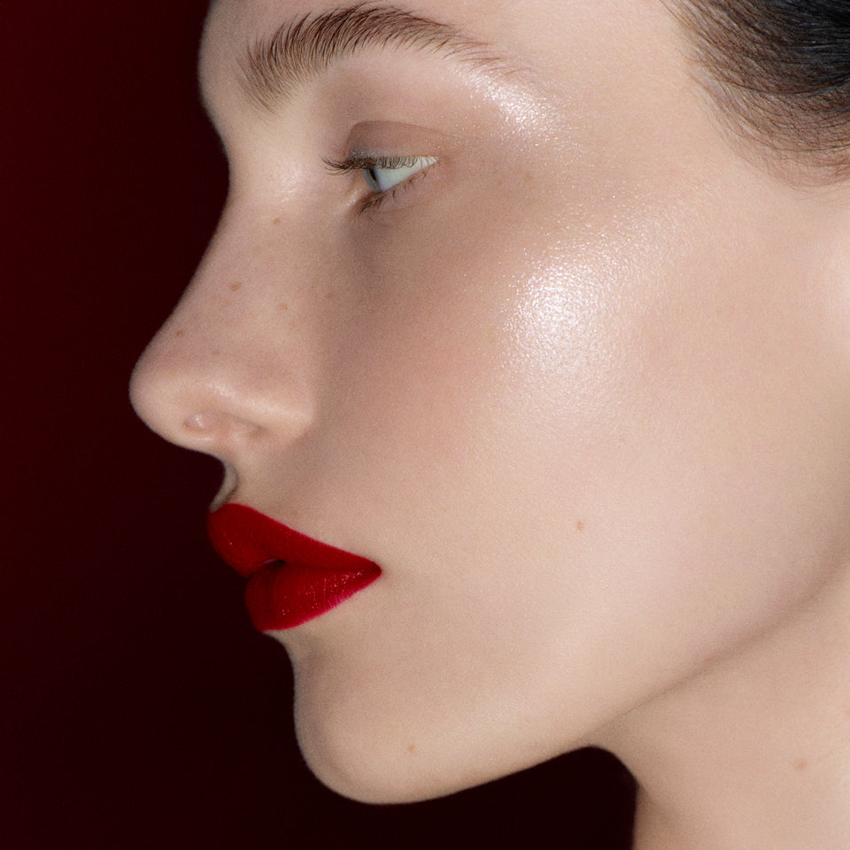 Profile of a fair-skinned model with luminous skin and a glossy red lip against deep burgundy — Victoria Beckham Beauty holiday collection.