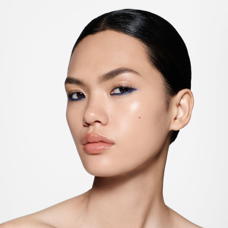 Close-up portrait of a model with dark hair pulled back, wearing blue eyeshadow and nude lipstick against a light gray studio background.