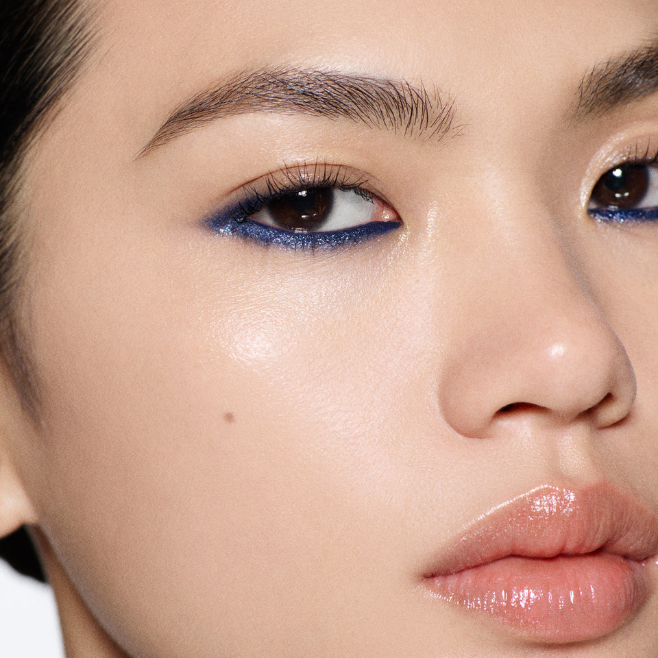 Close-up of a model's face showcasing a shimmering dark blue eyeshadow on the eyelid, paired with glossy nude-pink lips and neutral skin.