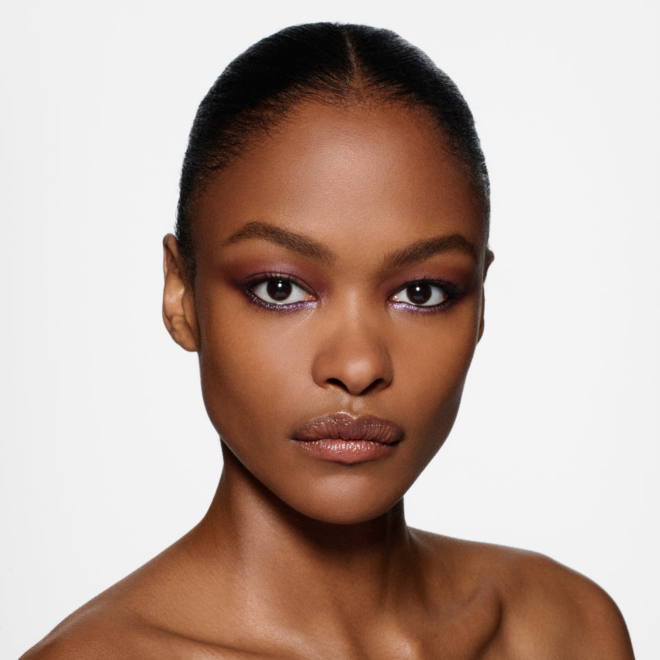 Deep-complexion model with slicked-back hair in a violet shimmer eye, facing forward — Victoria Beckham Beauty holiday look.