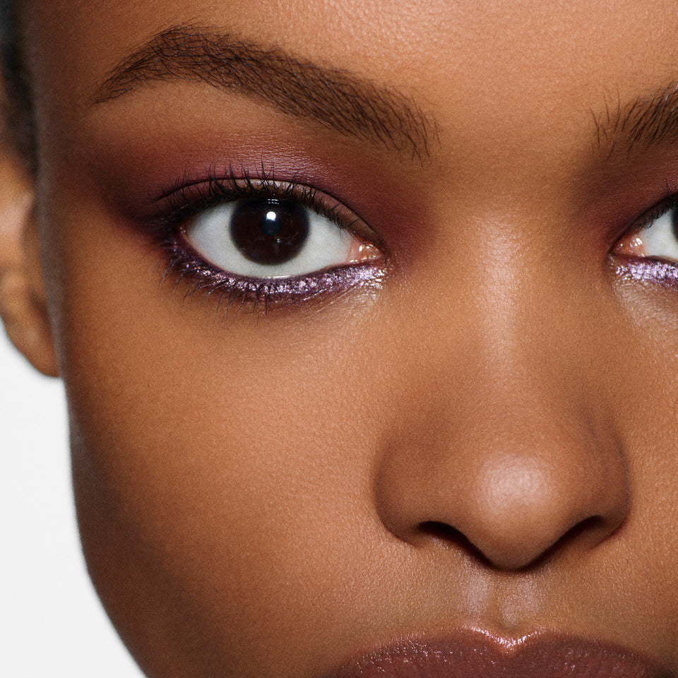 Close-up of a lilac shimmer eye and glossy lip on a deep-tan model — Victoria Beckham Beauty holiday eye look.