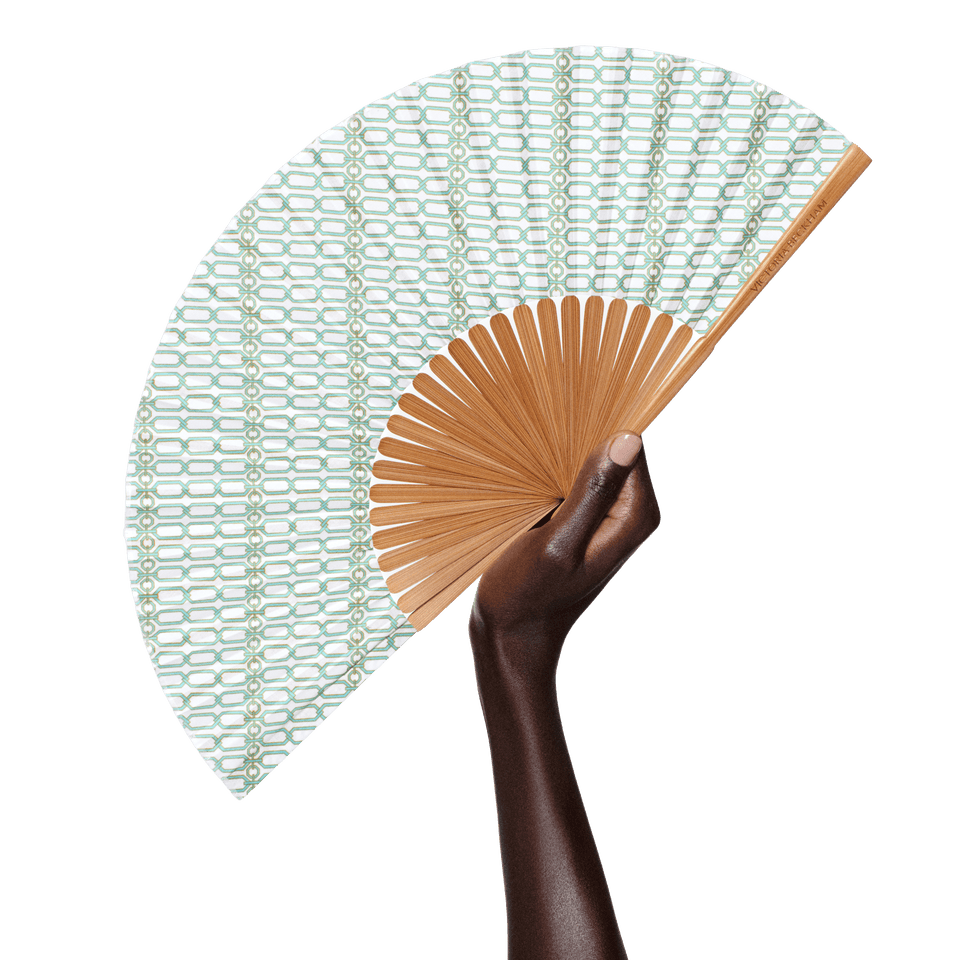 Deep-complexion hand raises an open Portofino '97 fan in turquoise and gold chain-link print by Victoria Beckham Beauty.