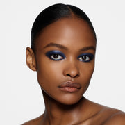 A close-up portrait of a model with slicked-back black hair, wearing dramatic blue eyeshadow and glossy nude lips against a clean white background.