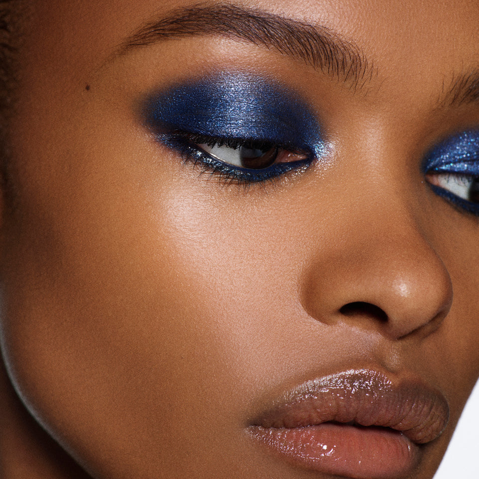 Close-up of a model's face showcasing a vibrant metallic blue eyeshadow look with shimmering blue eyeshadow on the eyelids and glossy nude-brown lips.
