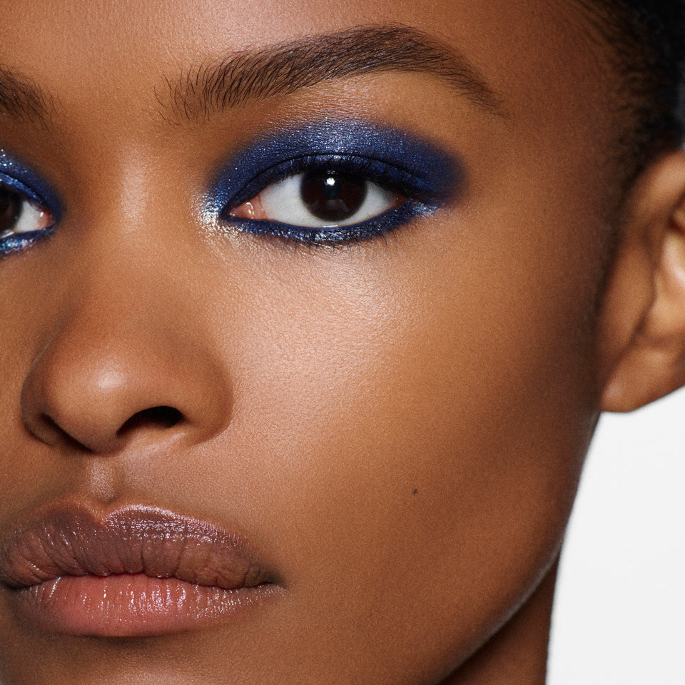 Close-up of a model's face showcasing a vibrant blue shimmery eyeshadow look with defined black winged eyeliner and neutral brown lipstick.