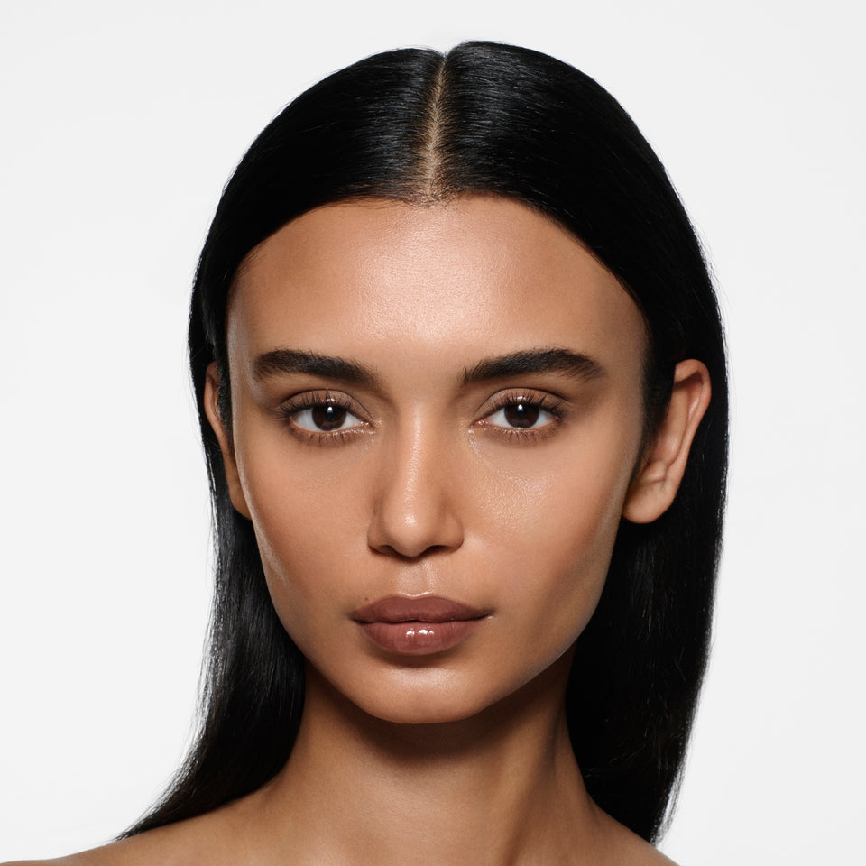 Tan-complexion model with long sleek centre-parted dark hair in Posh Gloss Poolside — cocoa brown by Victoria Beckham Beauty.