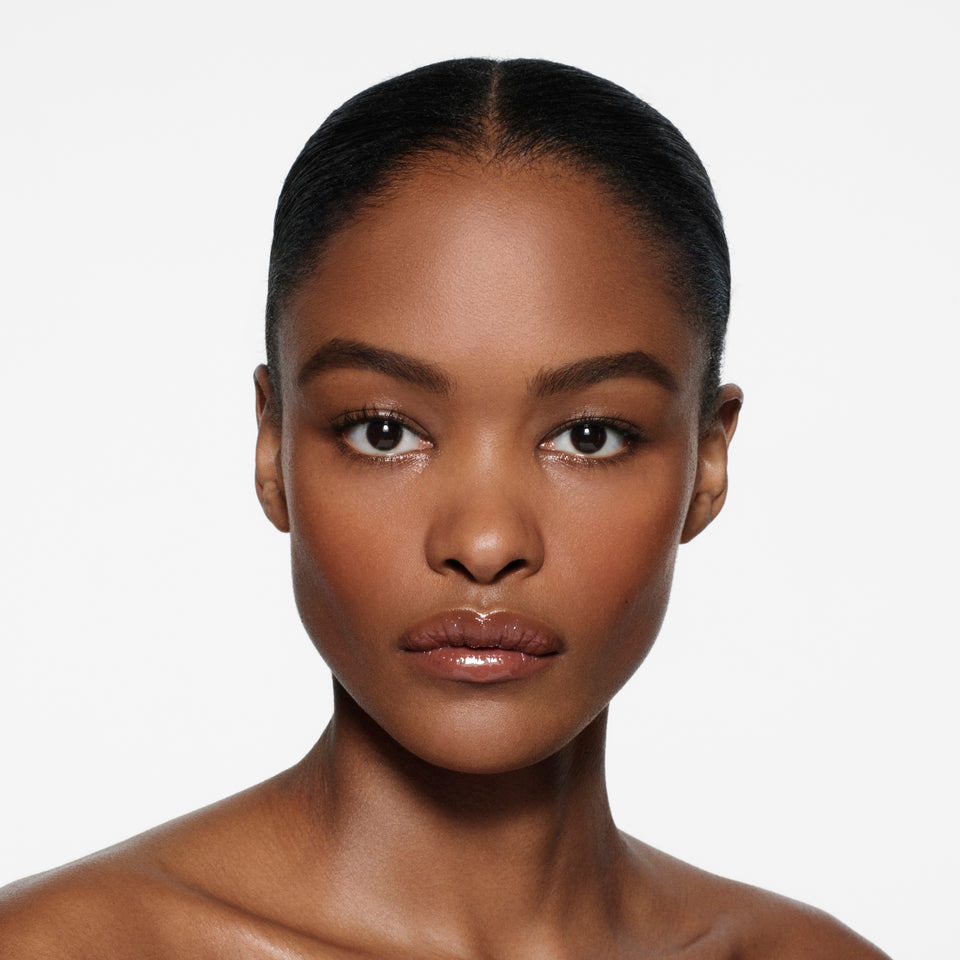 Brown-complexion model with slicked-back dark hair wearing Posh Gloss Poolside — cocoa brown by Victoria Beckham Beauty.