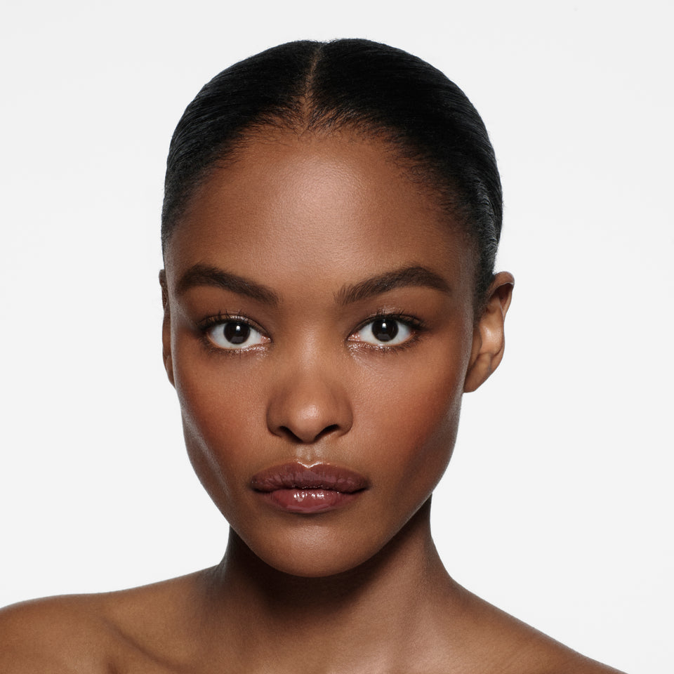 Brown-complexion model with sleek centre-parted dark hair wearing Posh Gloss Aperitivo — rosewood plum by Victoria Beckham Beauty.