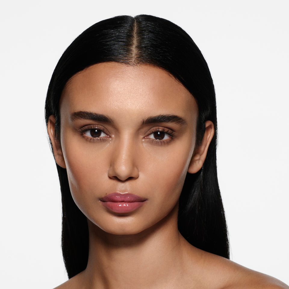 Tan-complexion model with long sleek dark hair wearing Posh Gloss Fizz — mauve berry by Victoria Beckham Beauty.
