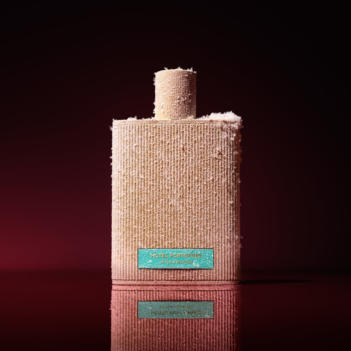 Snow-dusted ribbed Hotel Portofino bottle with turquoise label on a deep burgundy backdrop — Victoria Beckham Beauty for the holidays.