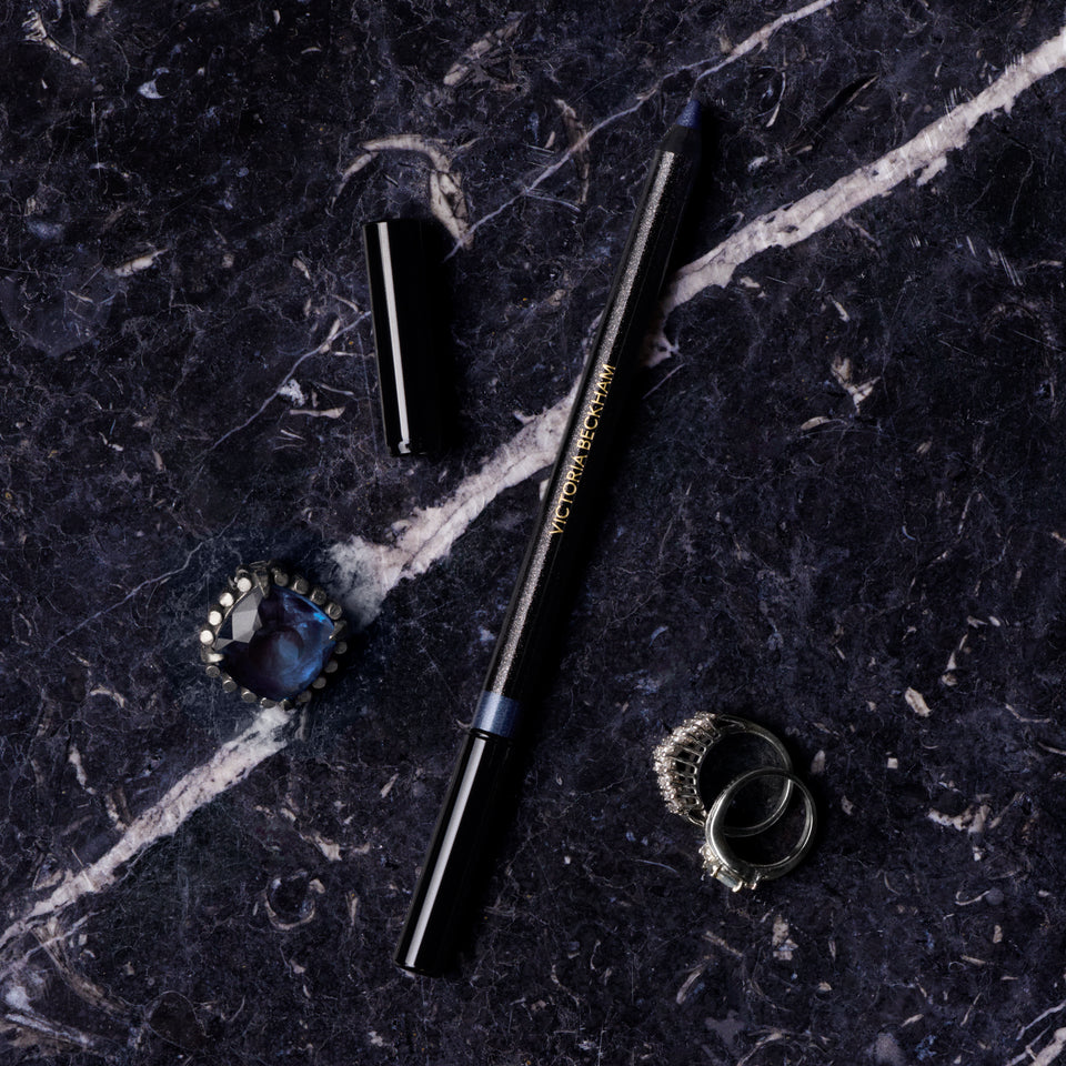 A black cosmetic pencil with gold lettering lies diagonally on a dark marble surface, alongside its cap, a blue gemstone ring, and a silver ring.