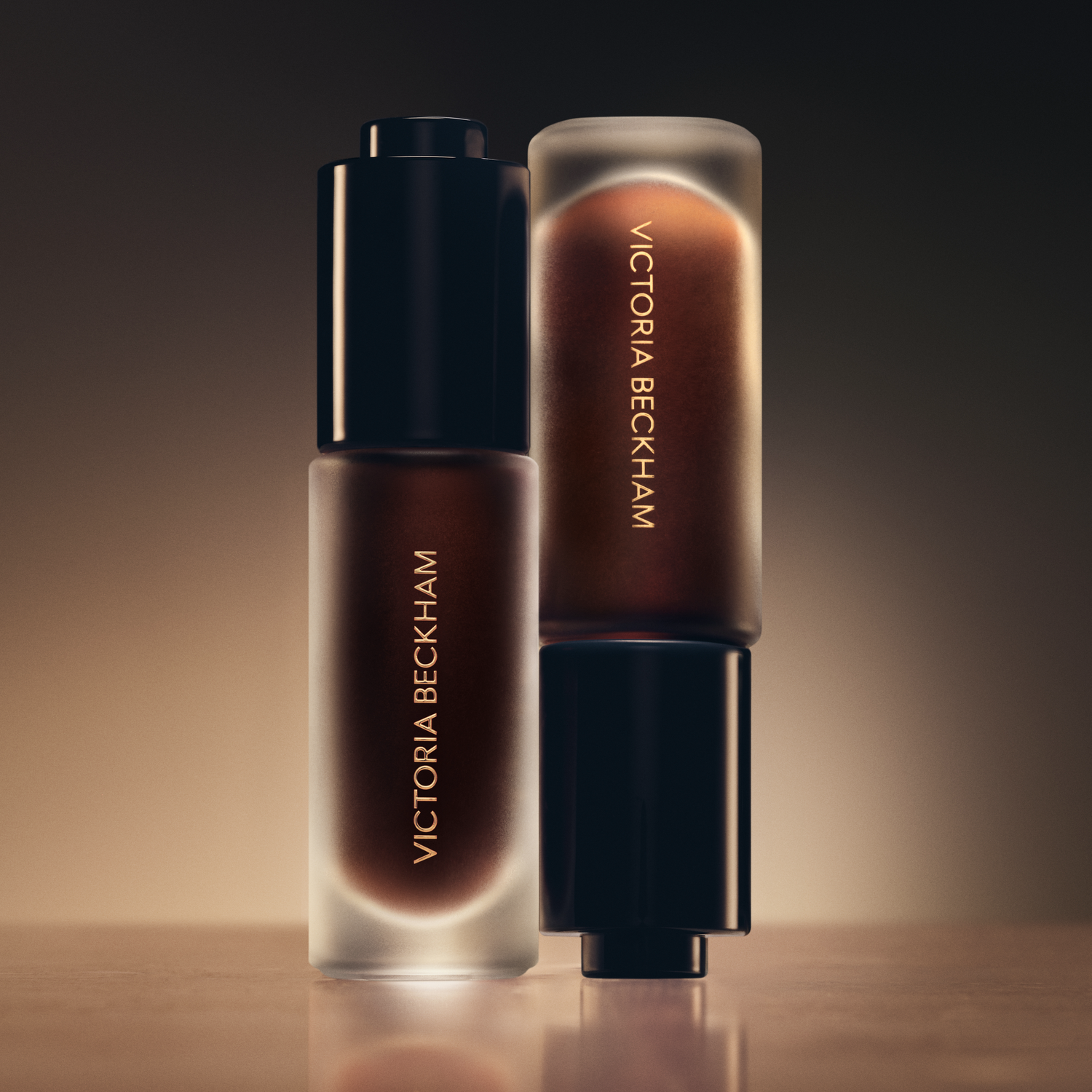Two Victoria Beckham Beauty product bottles spotlit against a warm amber backdrop — campaign still life.