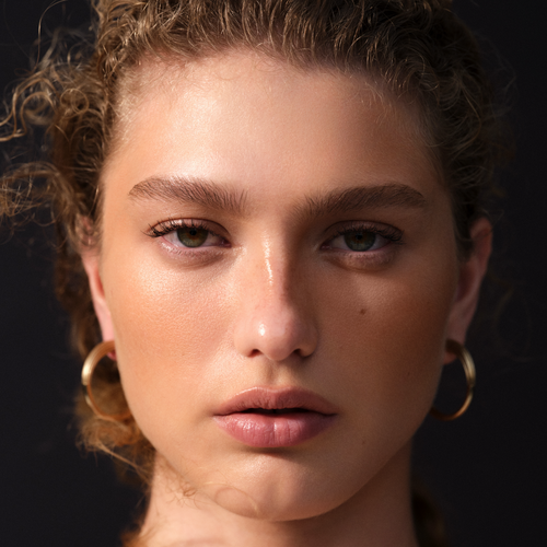 Curly-haired model in gold hoop earrings with glowing peach cheeks against a dark backdrop — Victoria Beckham Beauty campaign.