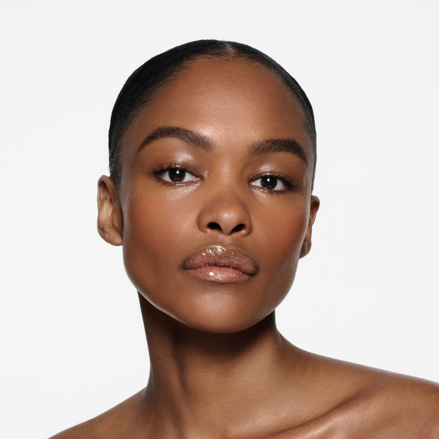Brown-complexion model with sleek slicked-back hair wearing Posh Gloss Martini — champagne gold shimmer by Victoria Beckham Beauty.