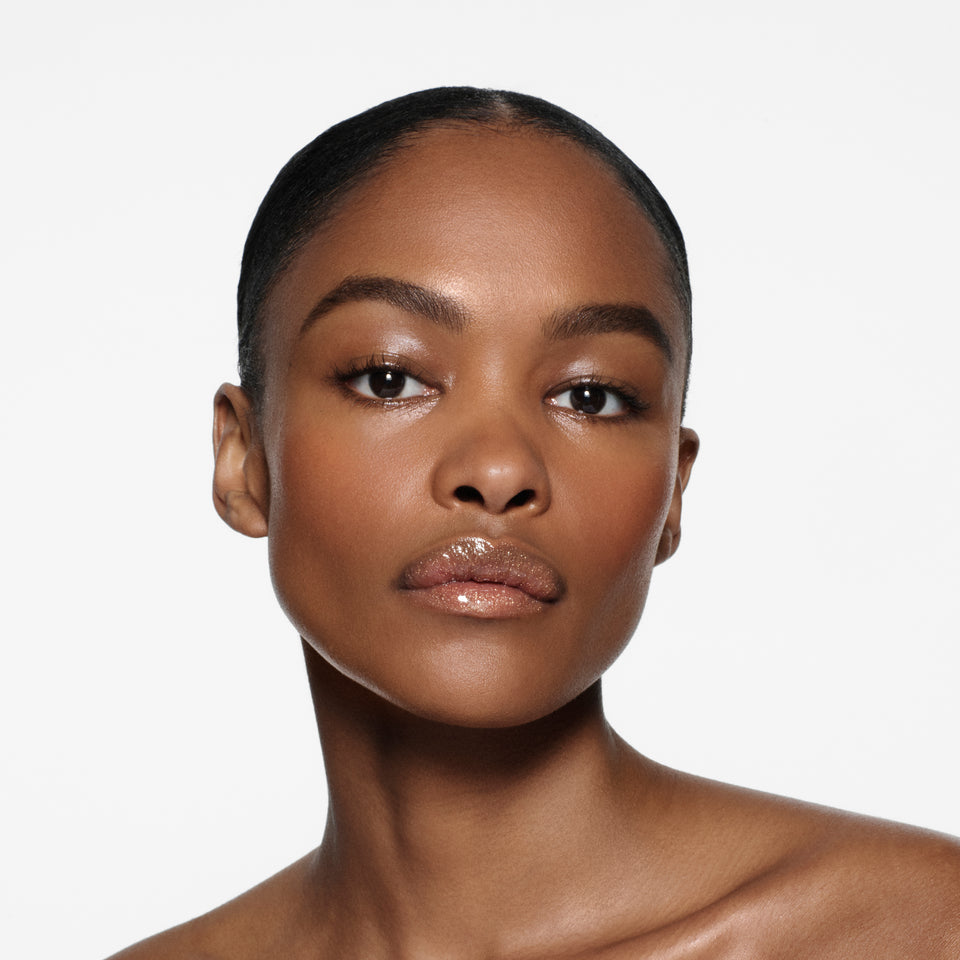 Brown-complexion model with sleek slicked-back hair wearing Posh Gloss Martini — champagne gold shimmer by Victoria Beckham Beauty.