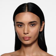 Olive-complexion model with long centre-parted dark hair wearing Posh Gloss Bungalow — warm coral peach by Victoria Beckham Beauty.