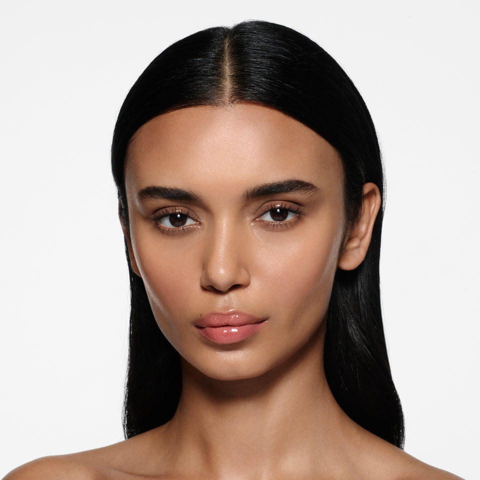 Olive-complexion model with long centre-parted dark hair wearing Posh Gloss Bungalow — warm coral peach by Victoria Beckham Beauty.