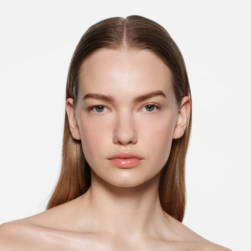 Fair-skinned model with sleek dirty-blonde hair in Posh Gloss Picante — vivid coral salmon by Victoria Beckham Beauty.