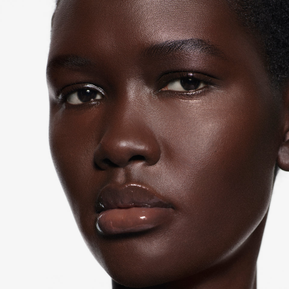 Close-up macro of lips on deep complexion glazed in Posh Gloss Ice — Victoria Beckham Beauty's clear mirror-finish lip gloss.