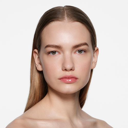Fair-skinned model with slicked-back light brown hair in Posh Gloss Bungalow — warm coral peach by Victoria Beckham Beauty.