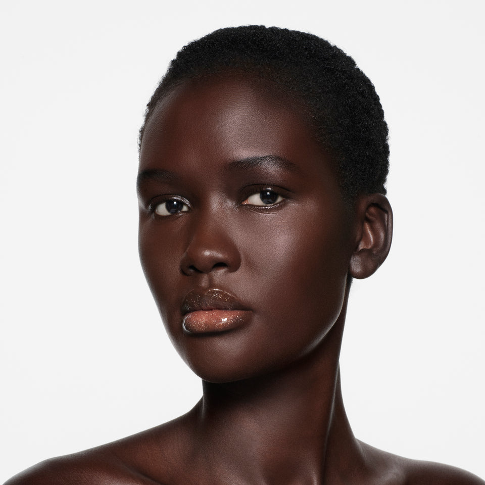 Deep-cocoa complexion with cropped natural hair, lips in Posh Gloss Martini — champagne gold shimmer by Victoria Beckham Beauty.