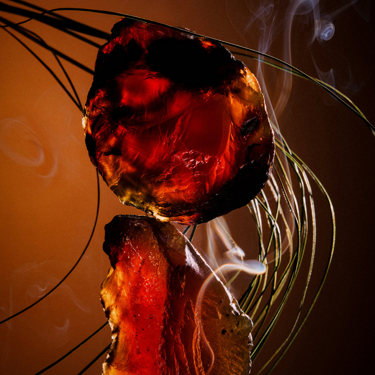 Backlit amber resin glowing red amid incense smoke and grasses — a black amber note in Hotel Portofino by Victoria Beckham Beauty.