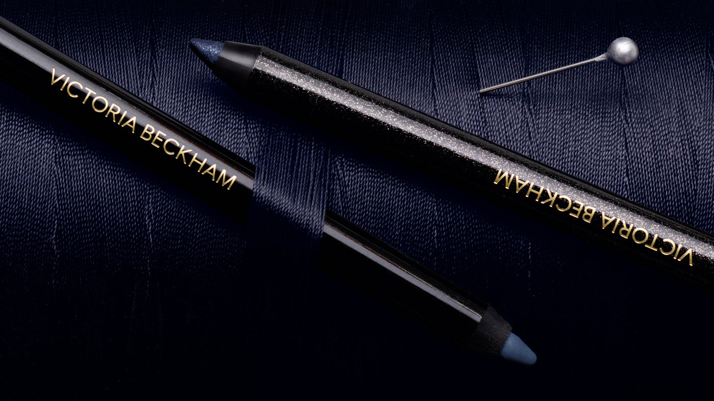 Two Victoria Beckham Satin Kajal Liners, one in glittering navy-silver and one in matte Navy Noir Pavé, laid diagonally across navy fabric with a decorative pin.