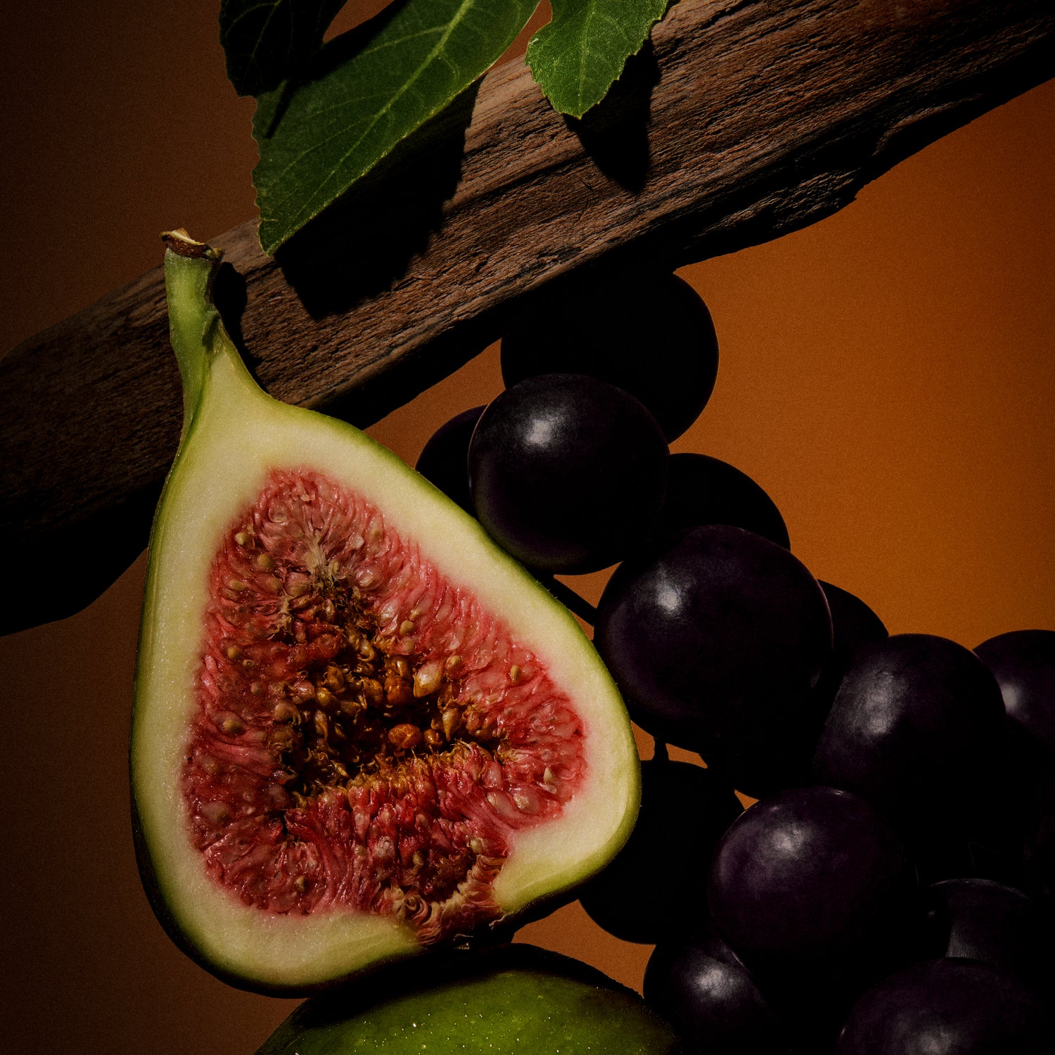Halved fig showing pink flesh beside dark grapes on a branch — wild fig in Hotel Portofino by Victoria Beckham Beauty.