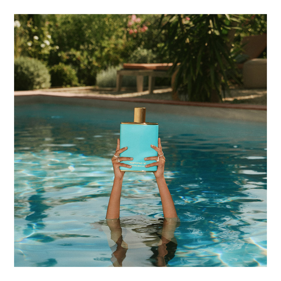 Two hands lift the gold-capped, turquoise Portofino '97 bottle from a sunlit garden pool at midday.