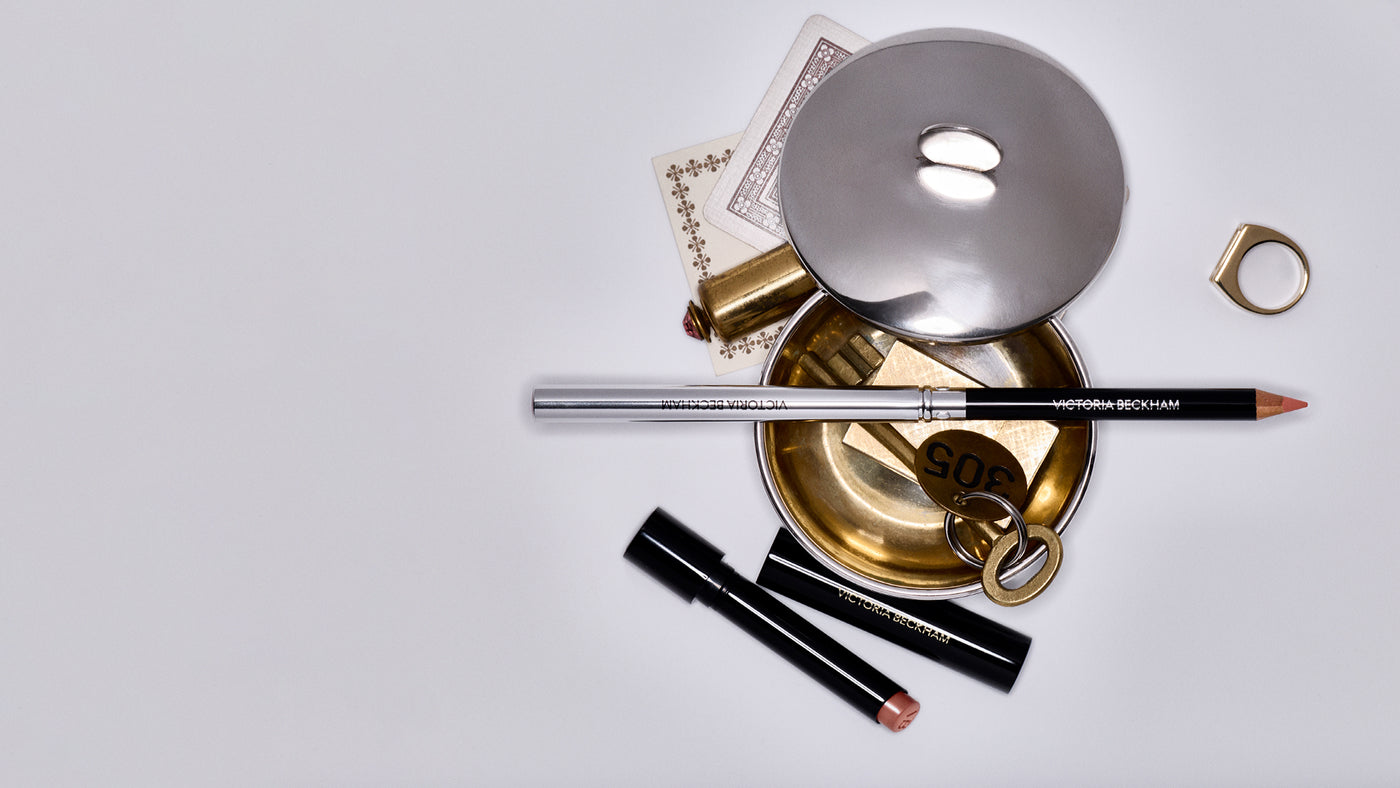 Victoria Beckham Beauty Lip Definer styled on a gold tray with cards, a ring and two Posh Lip bullets, desktop banner.
