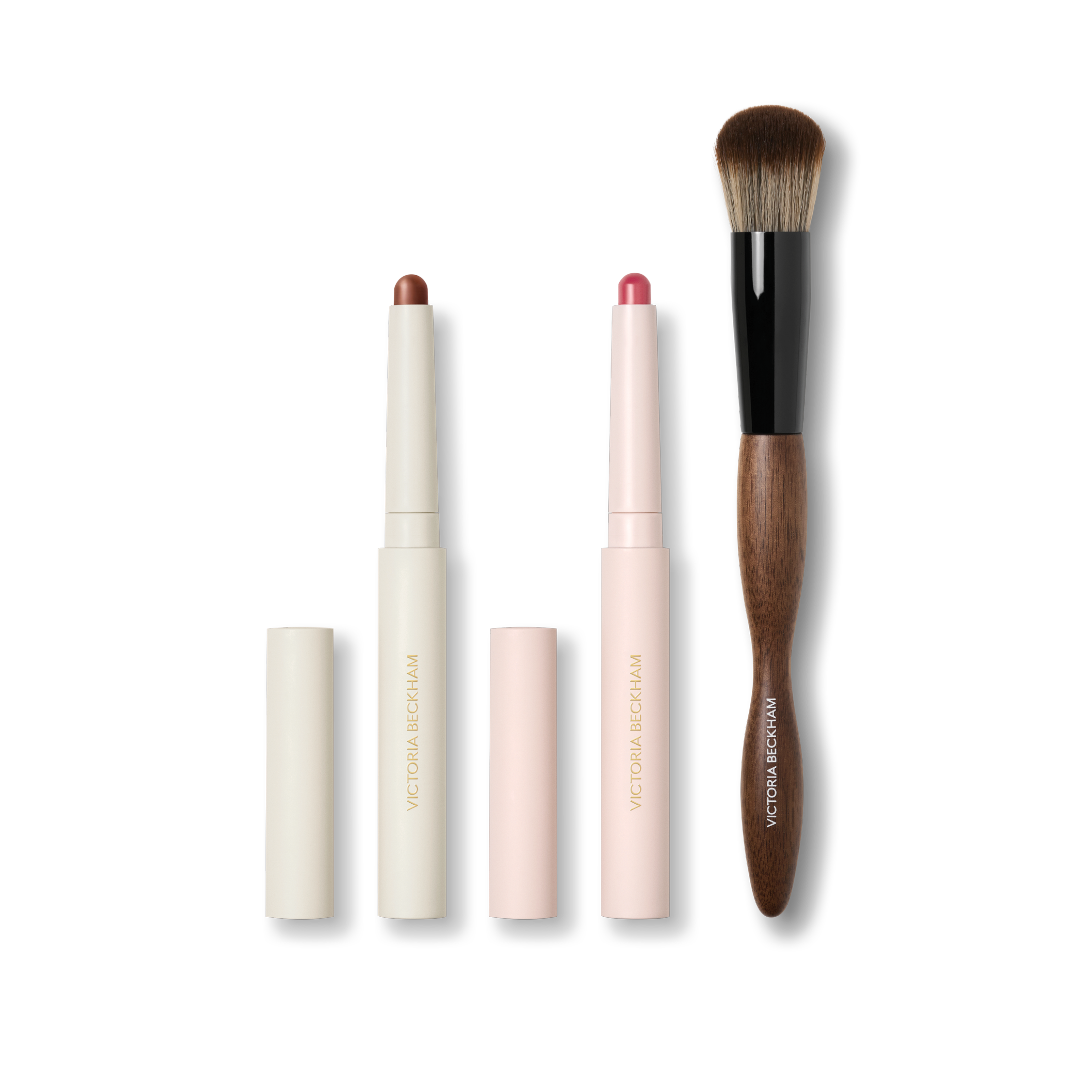 Cream Contour Stylus, pink Blush Stylus and a dual-ended wood-handled makeup brush on black — Victoria Beckham Beauty trio.