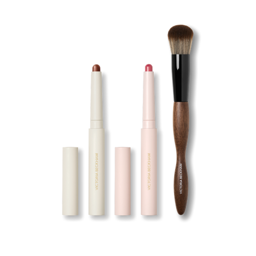 Cream Contour Stylus, pink Blush Stylus and a dual-ended wood-handled makeup brush on black — Victoria Beckham Beauty trio.