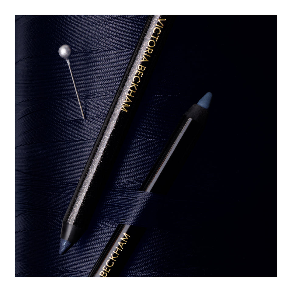 Victoria Beckham eyeliner pencil and matching pencil case on a dark navy textured fabric background.
