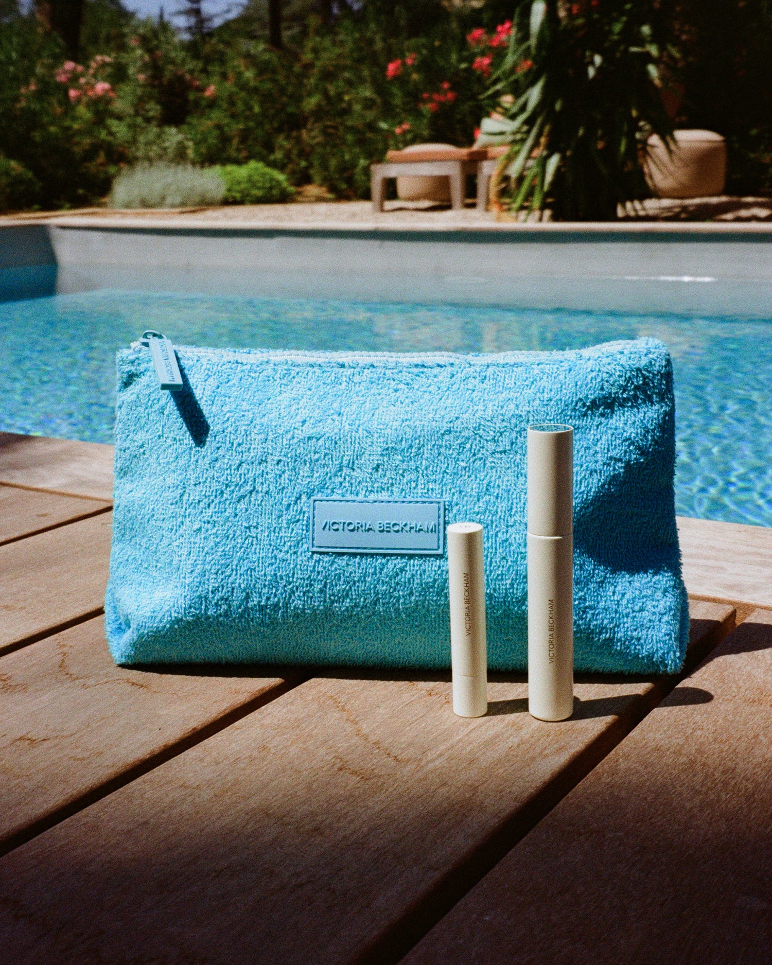 Closed Posh Balm Soleil and Vast Lash Cocoa beside a turquoise terry GWP pouch poolside — Victoria Beckham Beauty.