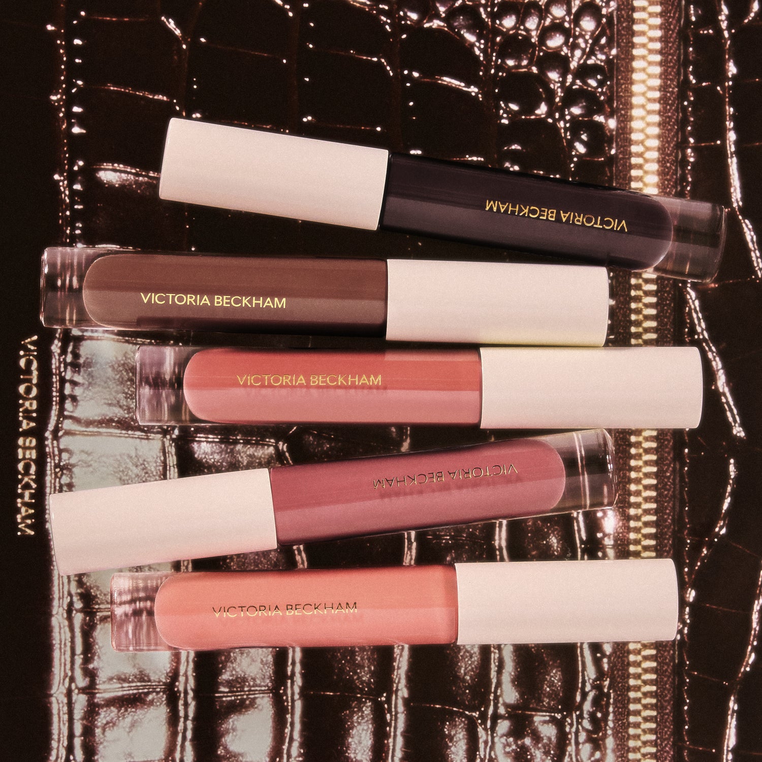 Five Posh Gloss tubes stacked on a brown crocodile clutch — Victoria Beckham Beauty's range of lip gloss shades.