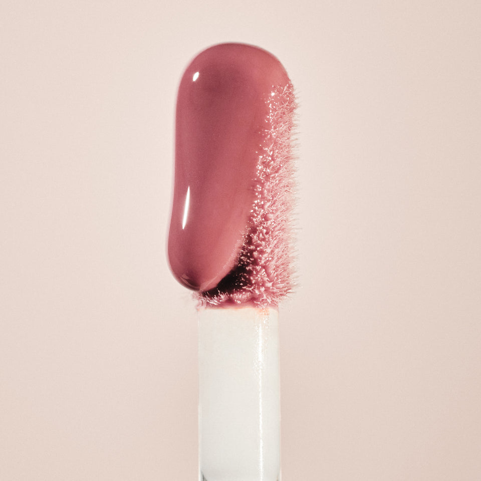 Macro of the Posh Gloss doe-foot wand laden with rosewood gloss against a blush backdrop — Victoria Beckham Beauty.