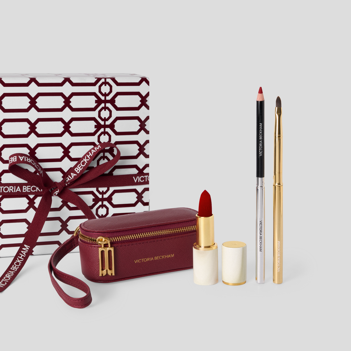 Victoria Beckham makeup gift set on a light gray background, featuring a burgundy zippered pouch, an open red lipstick, a black-and-silver pencil, a gold-and-silver pencil, and a patterned gift box with a matching ribbon.