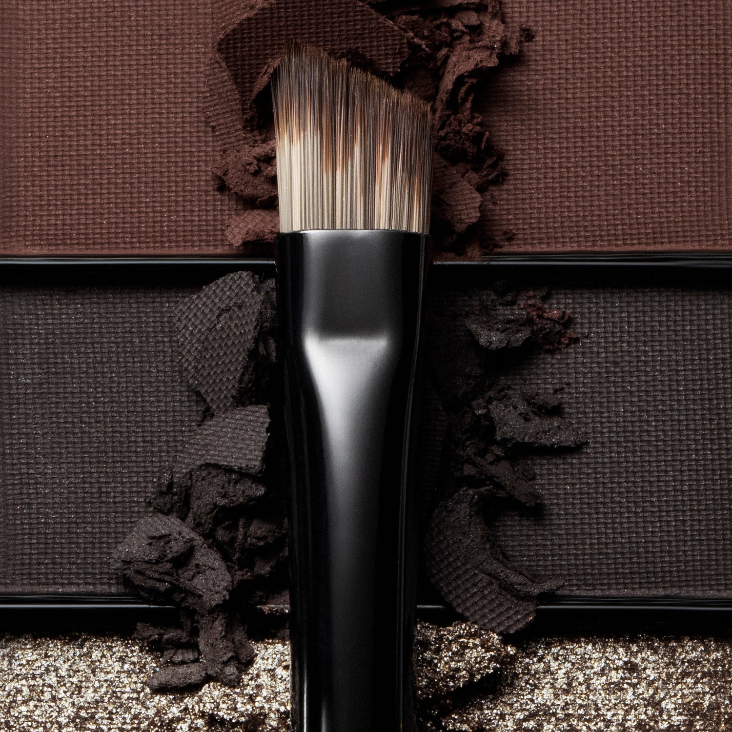 Close‑up of a makeup brush standing upright against crushed eyeshadow pans in deep brown, charcoal, and shimmering gold tones