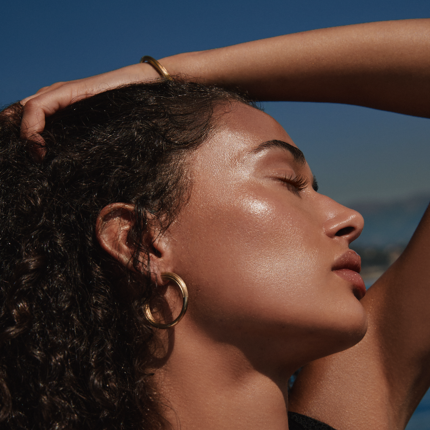 Sunlit profile close-up of a model with curly dark hair, gold hoop earrings and dewy skin, for Portofino '97.