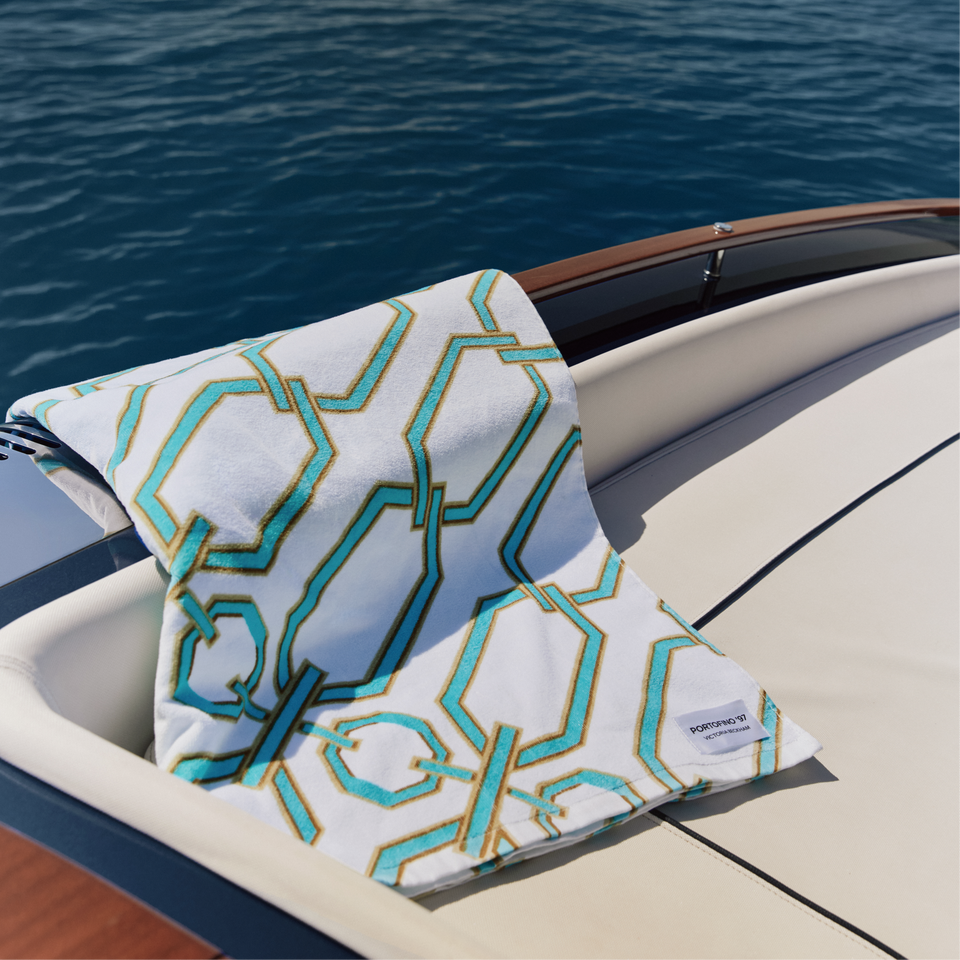 Turquoise and gold chain-link Portofino '97 towel draped over a speedboat's bow against open water.