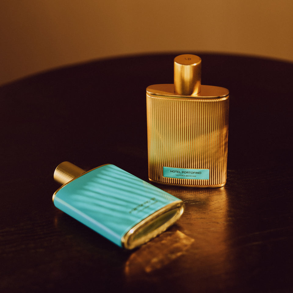 Two Hotel Portofino bottles, one fluted gold and one turquoise, on a dark wood table — the fragrance by Victoria Beckham Beauty.