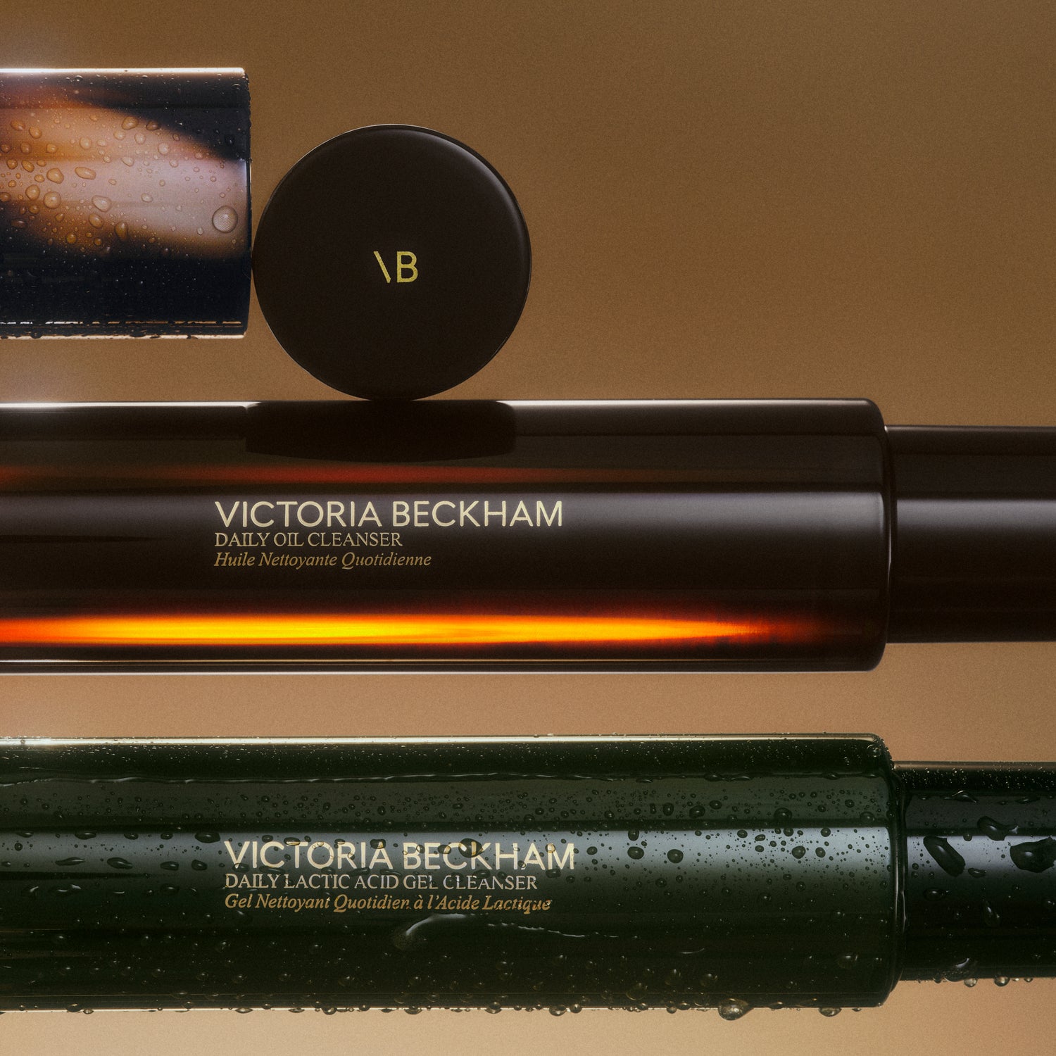 Victoria Beckham Beauty skincare bottles arranged horizontally, featuring the Daily Oil Cleanser and Daily Lactic Acid Gel Cleanser with water droplets on the packaging