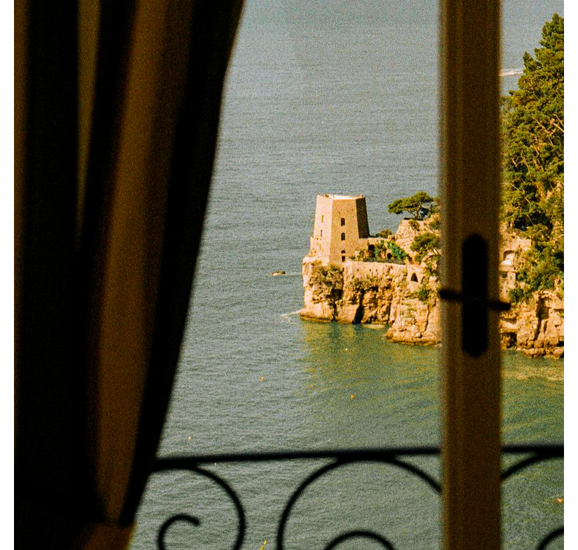 View through a shuttered window to a stone tower on a rocky Italian coastline — Hotel Portofino by Victoria Beckham Beauty.