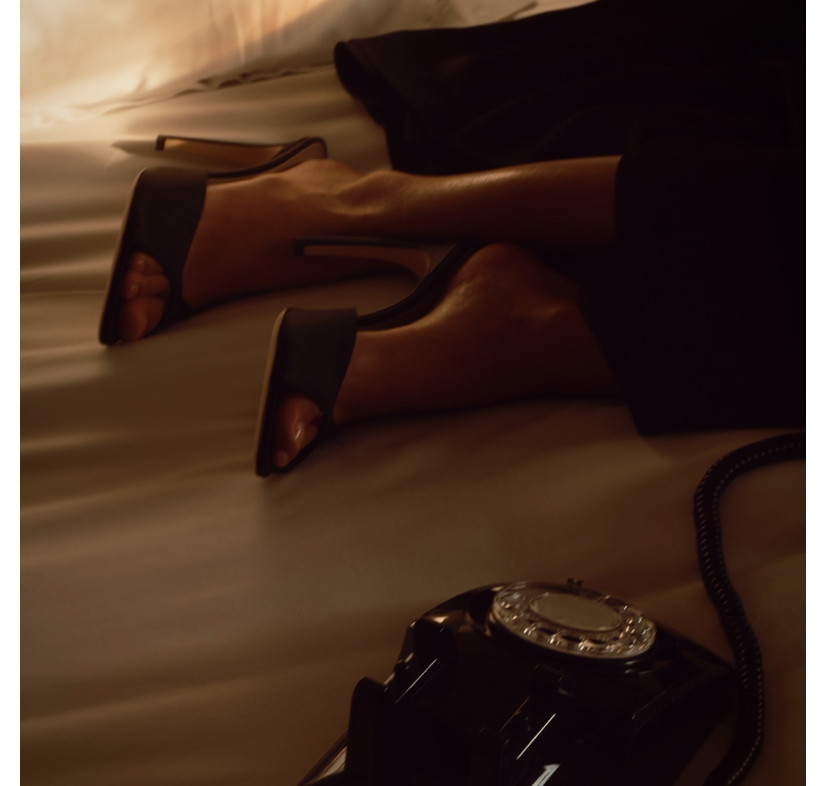 Legs in black satin mules resting on a cream bedspread beside a rotary telephone — Hotel Portofino by Victoria Beckham Beauty.