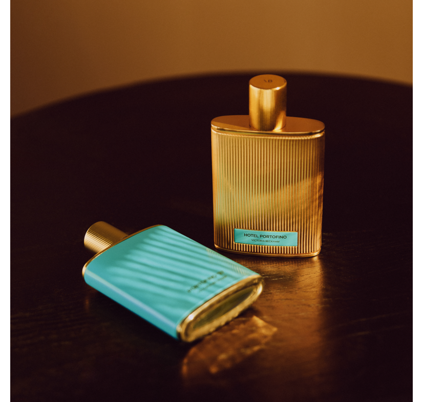 Two Hotel Portofino bottles, one fluted gold and one turquoise, on a dark wood table — the fragrance by Victoria Beckham Beauty.