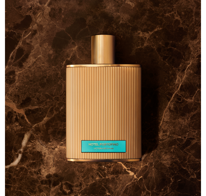 Fluted gold Hotel Portofino bottle with turquoise label on dark brown marble — the fragrance by Victoria Beckham Beauty.