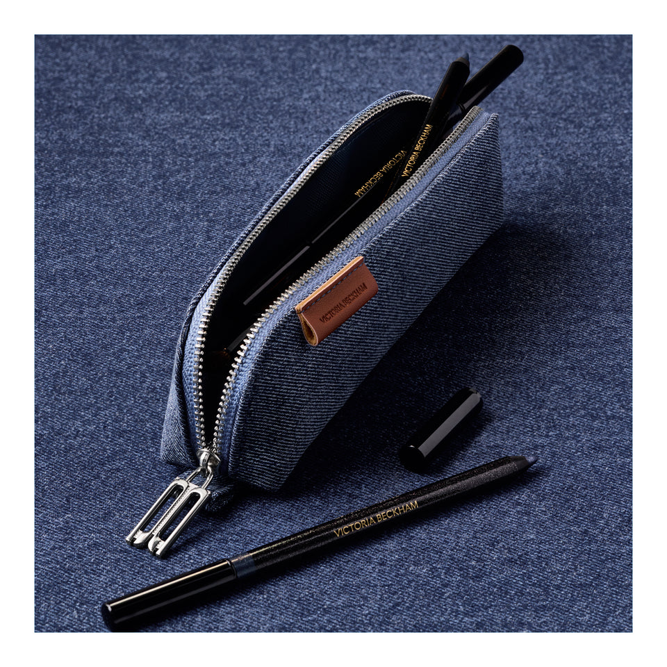 A blue-gray denim zippered pencil case shown open on a textured blue surface, with a black pencil and a small black cap placed outside it. 