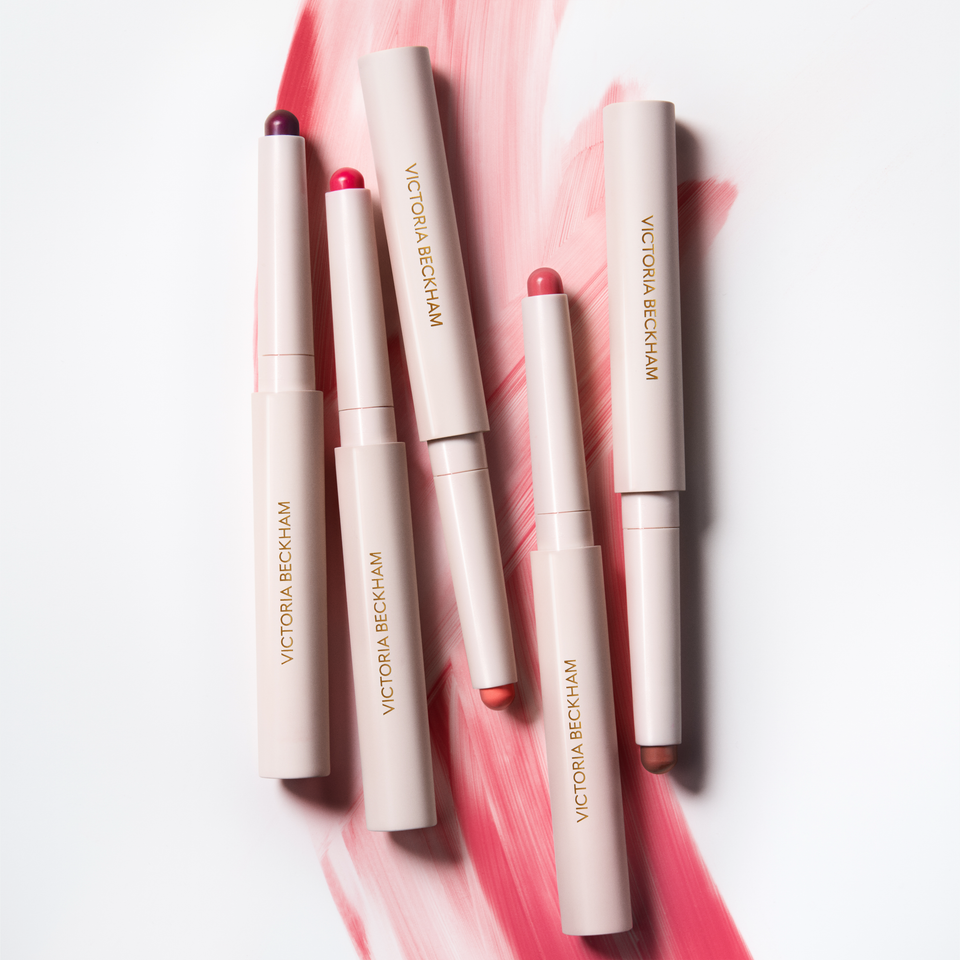 Five Blush Stylus pencils laid on a coral-pink brushstroke — Victoria Beckham Beauty's complete blush stick range.