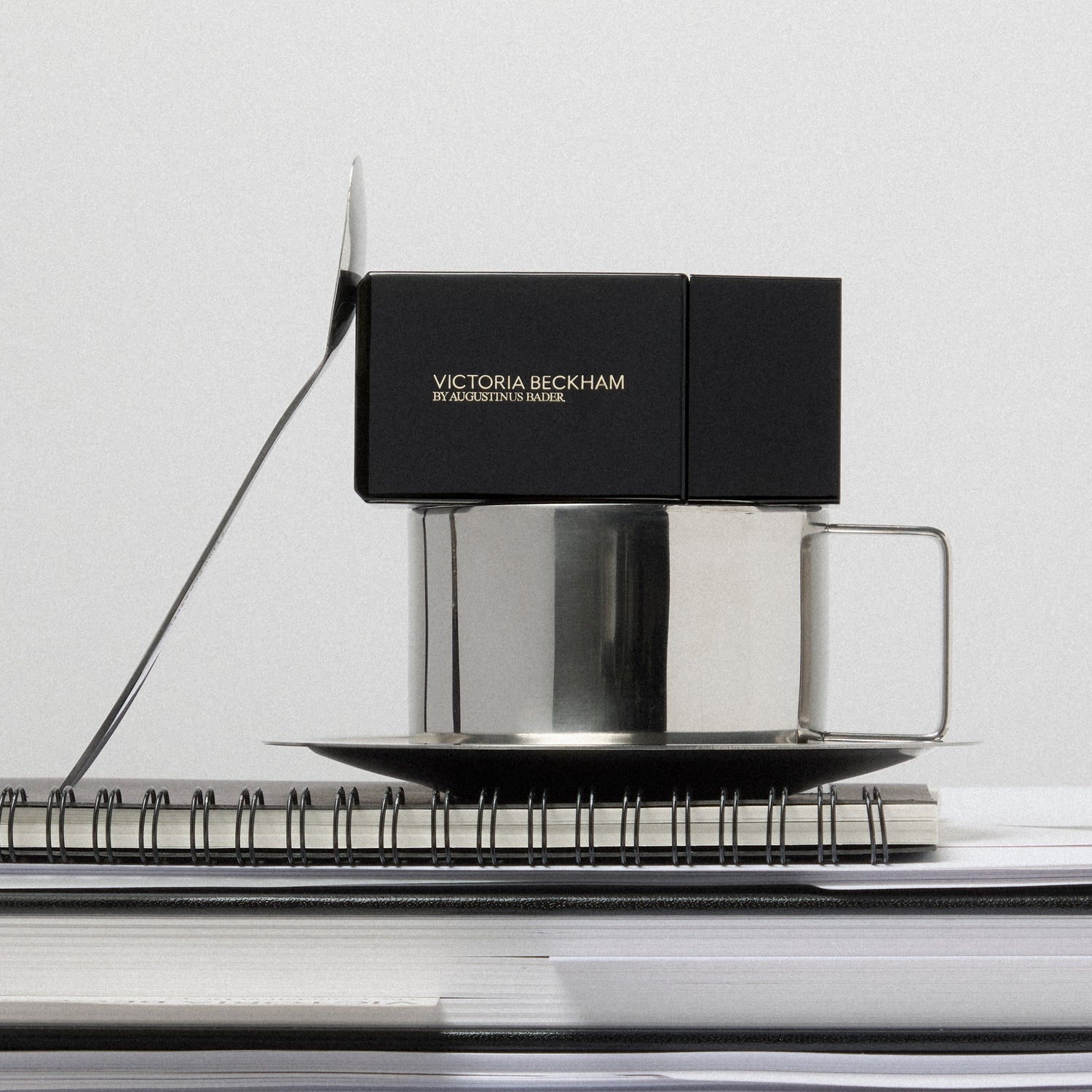 Boxed Victoria Beckham by Augustinus Bader Foundation Drops balanced on a silver cup and saucer atop a notebook.