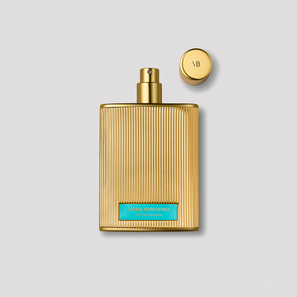 Fluted gold Hotel Portofino bottle uncapped beside its monogrammed cap on grey — the Extrait de Parfum by Victoria Beckham Beauty.
